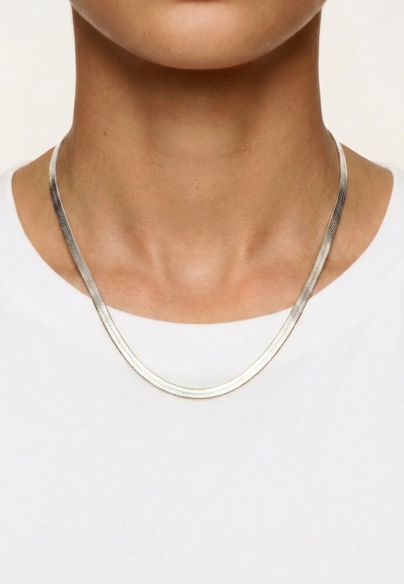 Collier chaîne en argent porté autour du cou d’une personne par-dessus une chemise blanche unie, vue rapprochée du menton jusqu’à la partie supérieure de la poitrine.