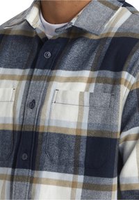 Flanellskjorta i marinblått, grått och krämfärgat rutigt mönster. Har en krage, två bröstfickor och knapfront med synlig sömnad.