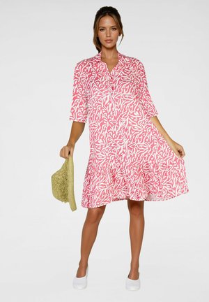 Žena v ružových a bielych šatách s abstraktným vzorom po kolená, drží slamák v jednej ruke a má na nohách biele nazúvacie topánky.