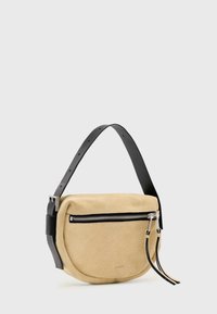 Sac demi-lune en daim beige avec sangle réglable en cuir noir, fermeture éclair argentée et deux tirettes en cuir noir.