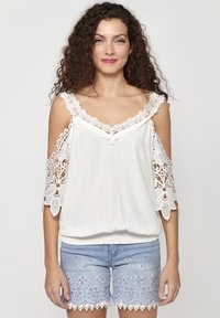 Blusa bianca con intricati accenti di pizzo sulle maniche scese, caratterizzata da una vestibilità ampia e orlo arricciato. Abbinata a pantaloni corti ricamati blu chiaro.
