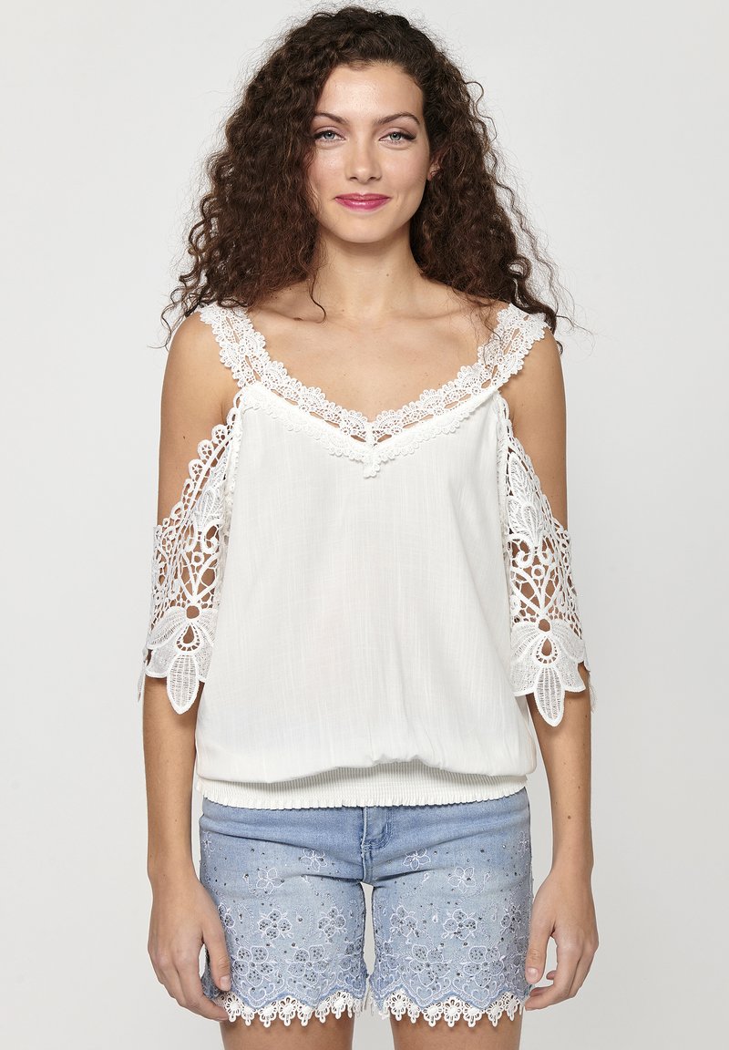 Blusa bianca con intricati accenti di pizzo sulle maniche scese, caratterizzata da una vestibilità ampia e orlo arricciato. Abbinata a pantaloni corti ricamati blu chiaro.
