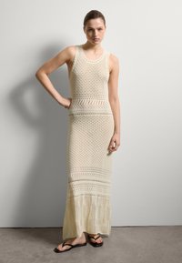 Strick, ärmelloses Maxikleid in Creme mit offenen Mustern, einer taillierten Oberseite und einem ausgestellten Saum. Schwarze Flip-Flops runden das Outfit ab.