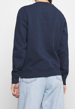 Person iført en navyblå langærmet sweatshirt og lyseblå jeans, vist bagfra mod en ensfarvet baggrund.