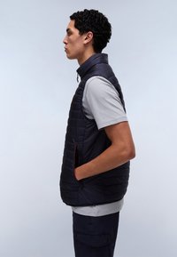 Muž v profile nosiaci čiernu prešívanú vestu na svetlo šedom tričku, ruky v vreckách vesty na jednoduchom svetlom pozadí.