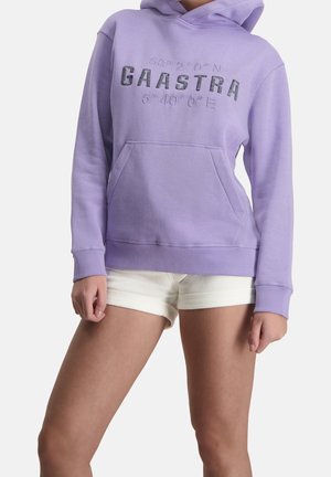 Lavendelfarbener Hoodie mit Fronttasche und schwarzem "GAASTRA"-Schriftzug, getragen mit weißen Shorts, zeigt Oberkörper und Beine vor einem einfarbigen Hintergrund.