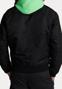 Svart bomberjacka med ribbat nederkant och ärmslut, med en grön huvtröja under, slät yta och utan synliga mönster.