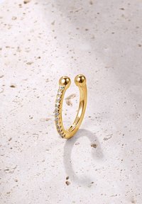 Goldring mit zwei sphärischen Enden, der eine Reihe kleiner, klarer Steine auf einer Seite aufweist. Glatte, polierte Oberfläche vor einem strukturierten Hintergrund.