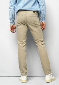Beige cotton trousers with a slim fit, featuring back pockets and a leather brand patch. Paired with white sneakers and a light blue shirt.