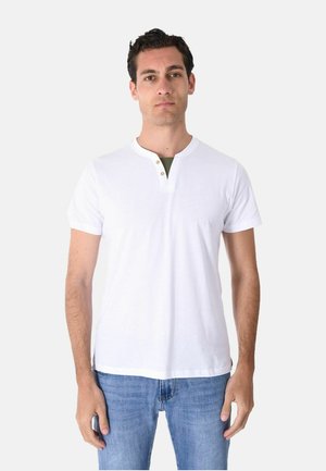 Uomo che indossa una maglietta henley bianca semplice a maniche corte e jeans blu, in piedi davanti a uno sfondo grigio chiaro.