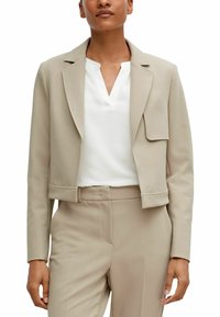 Beige kurze Jacke mit Revers, strukturierter Stoff und einer Brusttasche, getragen über einem weißen Hemd und kombiniert mit beigen Hosen.