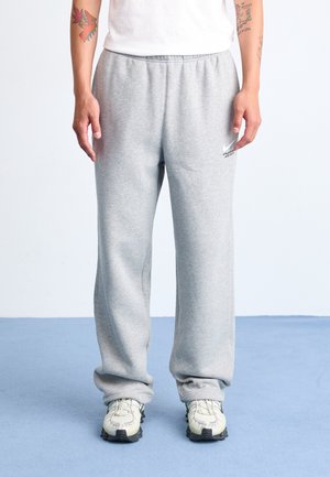 Personne portant un pantalon de jogging ample gris clair, des baskets blanches et une chemise blanche, debout sur un sol bleu avec des tatouages visibles sur les bras.