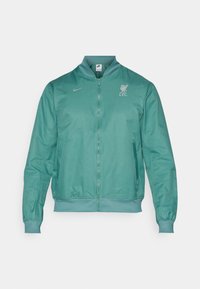 Teal bomberjakke lavet af glat stof. Forsiden har en fuld lynlås, ribbede manchetter og kant, samt et Liverpool FC-logo på brystet.