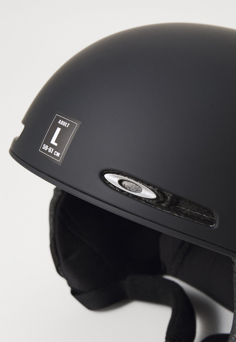 Svart vuxenhjälm med matt finish, storlek L (59-61 cm), har ett ventilationsöppning och ett varumärkets detalj. Mjuk innerslang synlig.