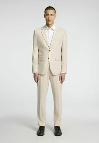 Jasnobeżowy garnitur z tkaniny tkanej, składający się z dopasowanej marynarki z dwoma guzikami, pasujących spodni oraz czarnych sznurowanych butów.