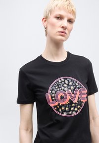 Άτομο με κοντά ξανθά μαλλιά που φοράει μαύρο μπλουζάκι με έναν κύκλο με σχέδιο λεοπάρδαλης και τη λέξη "LOVE" σε ροζ και πορτοκαλί γράμματα.