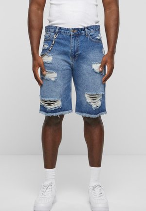 Shorts in denim con un lavaggio azzurro chiaro, caratterizzati da strappi consumati e orli sfrangiati, accompagnati da un accessorio a catena e indossati con sneaker bianche.