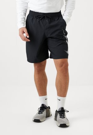 Homme portant un short noir avec cordon de serrage et un motif blanc sur le côté, des chaussettes blanches Nike, des chaussures de course grises et noires, et un haut blanc à manches longues.