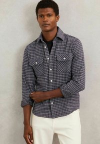 Cămașă în carouri, de culoare gri închis și alb, realizată dintr-un material texturat. Dispune de un guler, două buzunare pe piept și închidere cu nasturi în față. Mânecile sunt rulate.