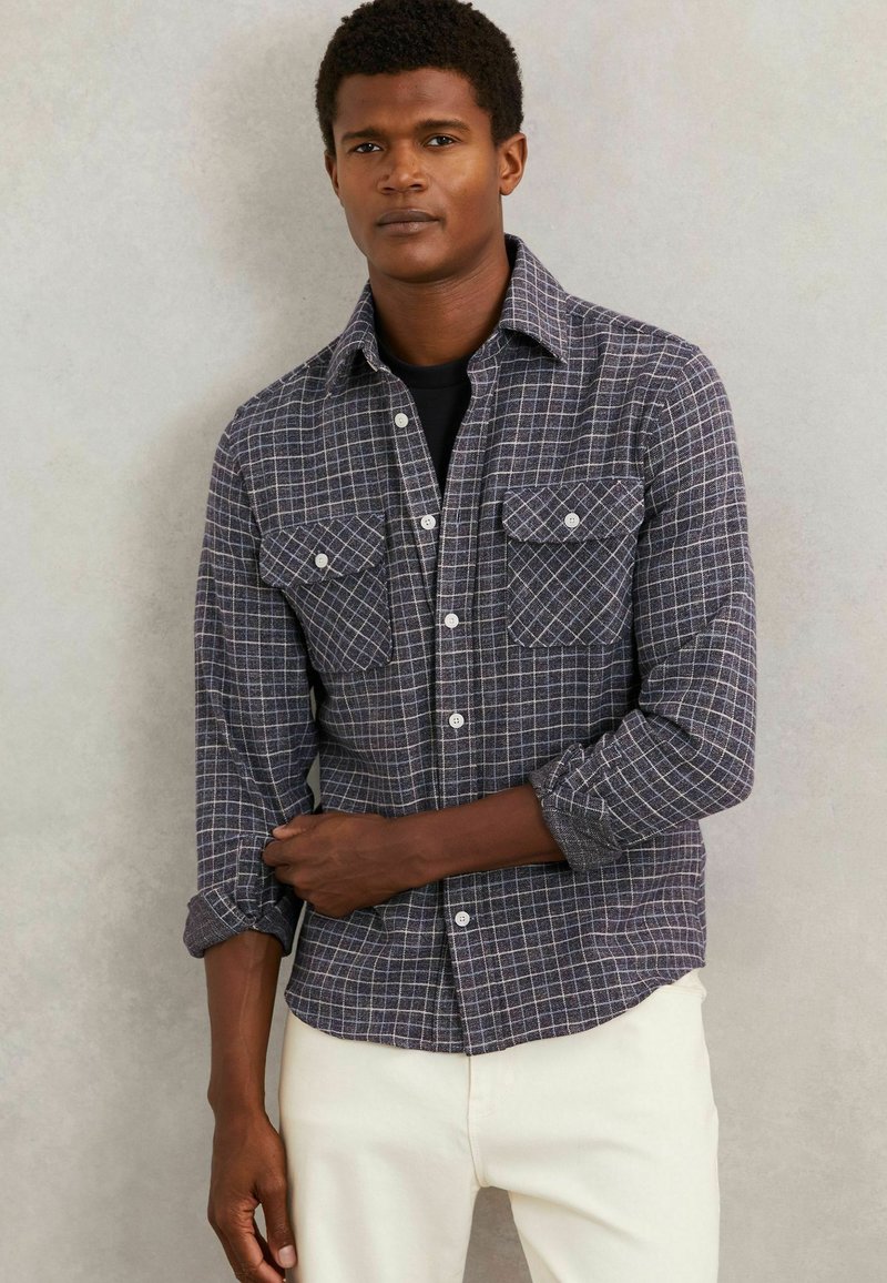 Cămașă în carouri, de culoare gri închis și alb, realizată dintr-un material texturat. Dispune de un guler, două buzunare pe piept și închidere cu nasturi în față. Mânecile sunt rulate.