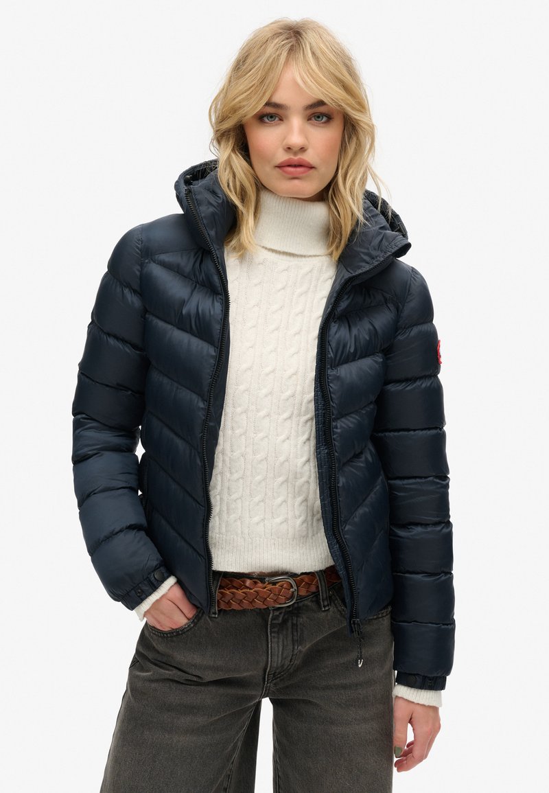 Marineblauwe pufferjack met horizontaal gestikt ontwerp, ritssluiting en capuchon. Gedragen over een crèmekleurige kabeltrui en grijze spijkerbroek.