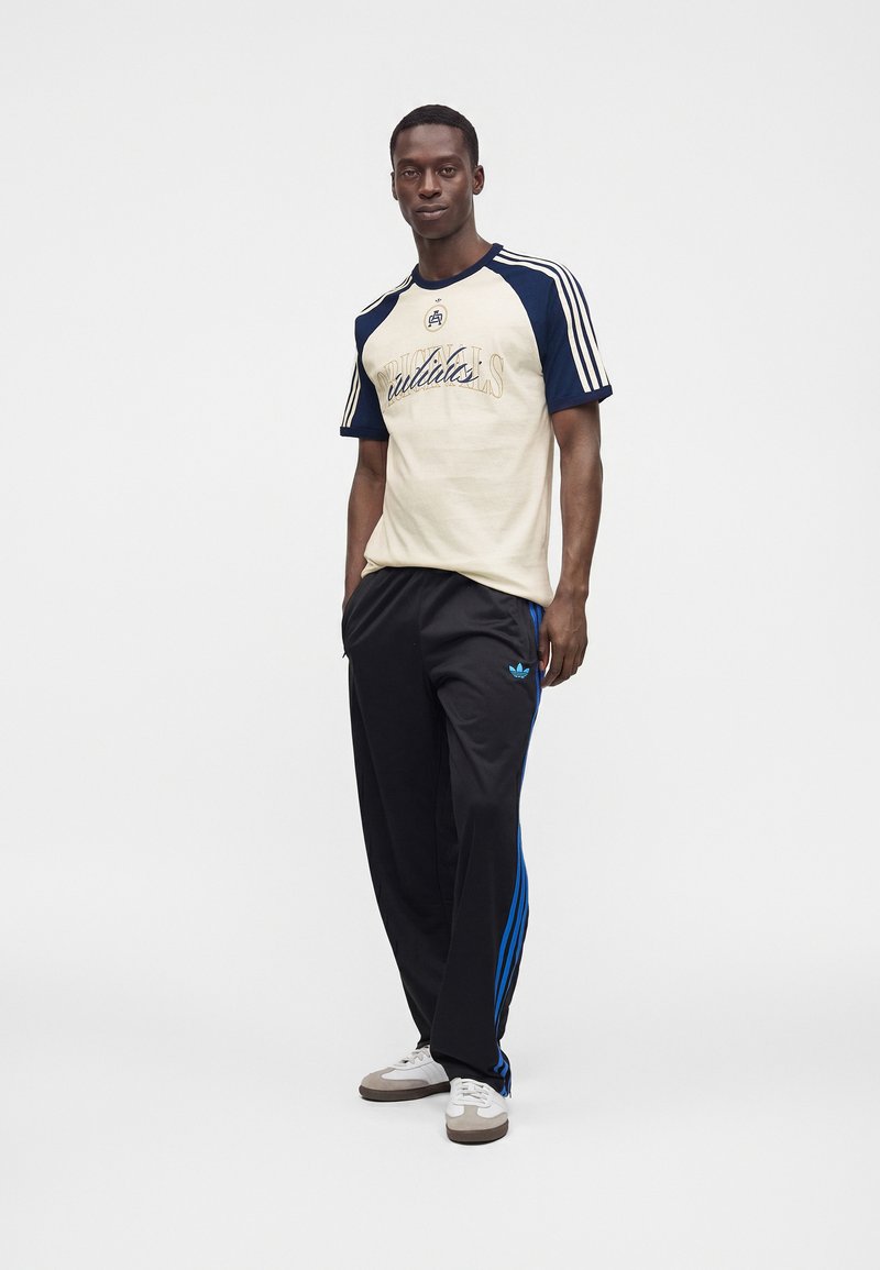 Άνδρας που φοράει μπλουζάκι Adidas κοντομάνικο μπεζ και ναυτικό, μαύρη φόρμα Adidas με μπλε ρίγες και λευκά αθλητικά παπούτσια, στέκεται μπροστά σε λευκό φόντο.