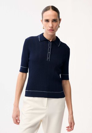 Femme aux cheveux coiffés en arrière portant un haut en maille bleu marine à manches courtes avec bordure blanche et un pantalon blanc taille haute, debout devant un fond uni.