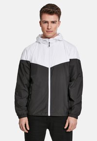 Veste coupe-vent noir et blanc avec capuche, fermeture �éclair frontale et design coloré avec texture lisse et poignets élastiques.