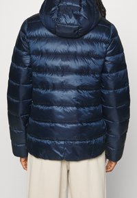 En person som bär en marinblå vadderad jacka med huva, sedd från baksidan, tillsammans med ljusbeiga byxor.