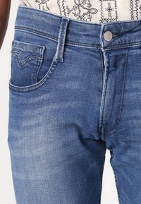 Gros plan d'un jean en denim bleu moyen avec bouton devant, fermeture éclair, passants de ceinture et poche avant sur une chemise blanche à motifs.