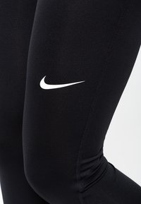 Zbliżenie czarnych legginsów sportowych z białym logo Nike na udzie oraz widocznymi detalami szycia.