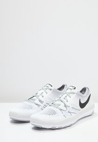 Białe buty sportowe z przewiewną siateczką u góry, szarymi akcentami i czarnym logo Nike w kształcie „swoosh”. Posiadają elastyczną podeszwę i system sznurowania.