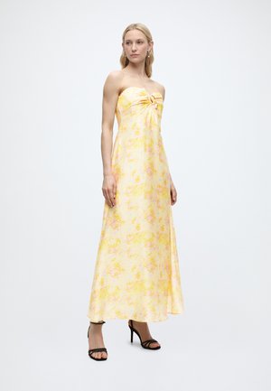 Γυναίκα όρθια, φοράει μάξι φόρεμα χωρίς τιράντες με κίτρινα λουλούδια και λεπτομέρεια κόμβου, συνδυασμένο με μαύρα ψηλοτάκουνα σανδάλια, απλό λευκό φόντο.