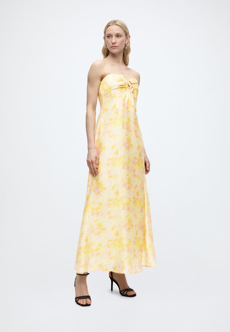 Γυναίκα όρθια, φοράει μάξι φόρεμα χωρίς τιράντες με κίτρινα λουλούδια και λεπτομέρεια κόμβου, συνδυασμένο με μαύρα ψηλοτάκουνα σανδάλια, απλό λευκό φόντο.