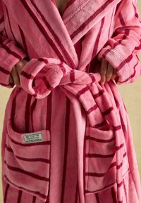 Różowy pluszowy szlafrok z pionowymi czerwonymi paskami, zapinany nawiązaniem, z dwiema przednimi kieszeniami i związanym paskiem. Miękka, futrzasta tekstura.