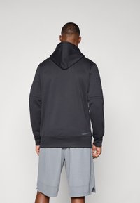 Juoda hooded sweatshirt su lygiu paviršiumi, turintis aukštą apykaklę ir banguotas rankogalius, derinamas su pilkais sportiniais šortais.