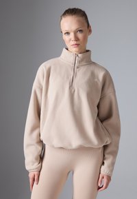 Tânără purtând un pulover bej din fleece cu fermoar pe jumătate și colanți cu nervuri asortați, stând în fața unui fundal gri simplu.