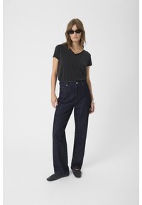 Black v-neck t-shirt, high-waisted dark denim jeans, and black flat shoes. Model wears sunglasses, standing on a plain background.