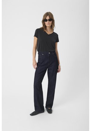 Black v-neck t-shirt, high-waisted dark denim jeans, and black flat shoes. Model wears sunglasses, standing on a plain background.