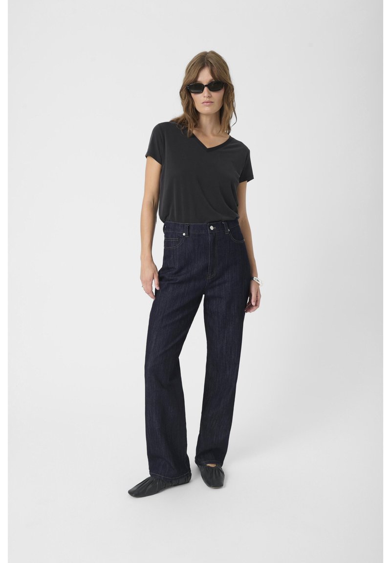 Black v-neck t-shirt, high-waisted dark denim jeans, and black flat shoes. Model wears sunglasses, standing on a plain background.