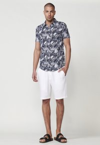 Kratka majica s kratkimi rokavi v mornarsko modri barvi z belim listnatim vzorcem, v kombinaciji z belimi kratkimi hlačami in črnimi sandali. Ima gumbe in sproščen kroj.