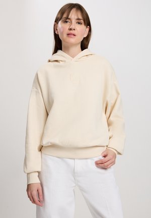 Kremowy, oversize'owy hoodie z kieszenią kangurką z przodu, ściągaczami na rękawach oraz subtelnym wyszytym logo na klatce piersiowej. Połączony z białymi spodniami.