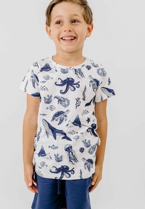 Lächelnder Junge mit weißem T-Shirt mit blauen Meeresbewohnern und dunkelblauen Shorts, der vor einem einfarbigen hellen Hintergrund steht.