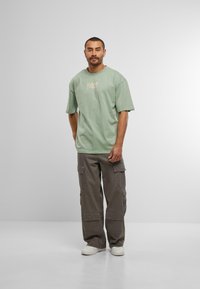 Lys grøn oversized t-skjorte med logo foran, kombinert med grå cargo-bukser med flere lommer og en avslappet passform. Hvite sneakers.