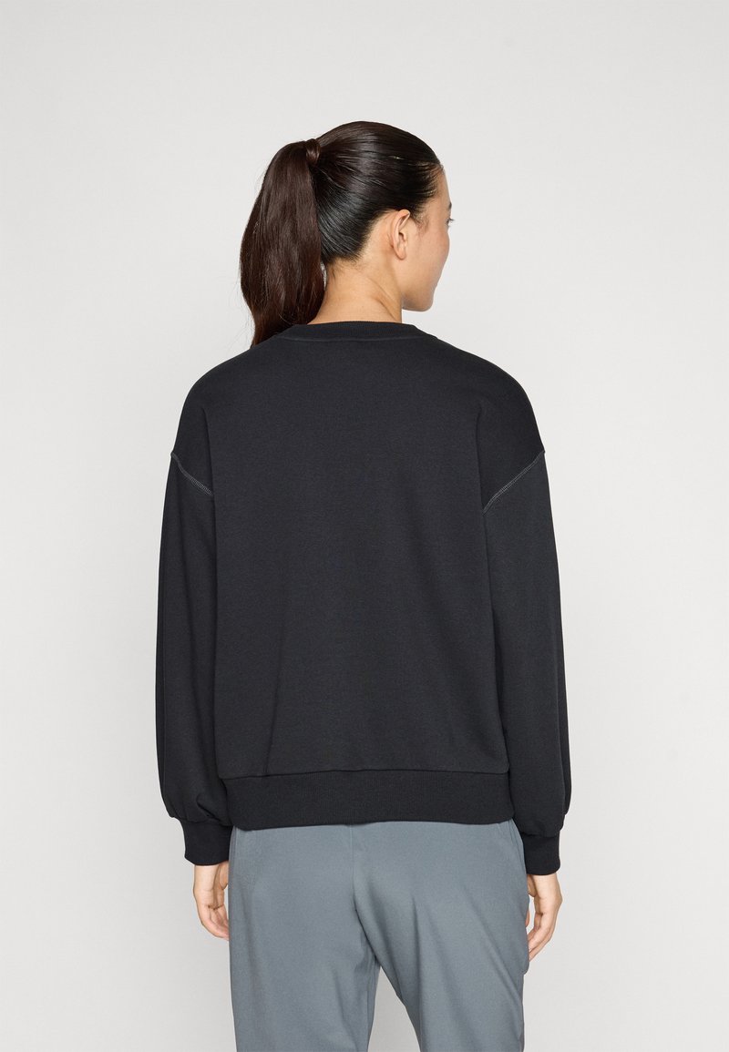 Felpa nera con maniche lunghe e voluminose, scollatura a girocollo e polsini a coste. Il materiale sembra morbido e liscio. Indossata con pantaloni grigi.