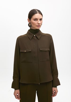 Blusa marrone a maniche lunghe con colletto alto, due tasche frontali, dettagli in bottone color oro e tessuto testurizzato, abbinata a un dolcevita coordinato.
