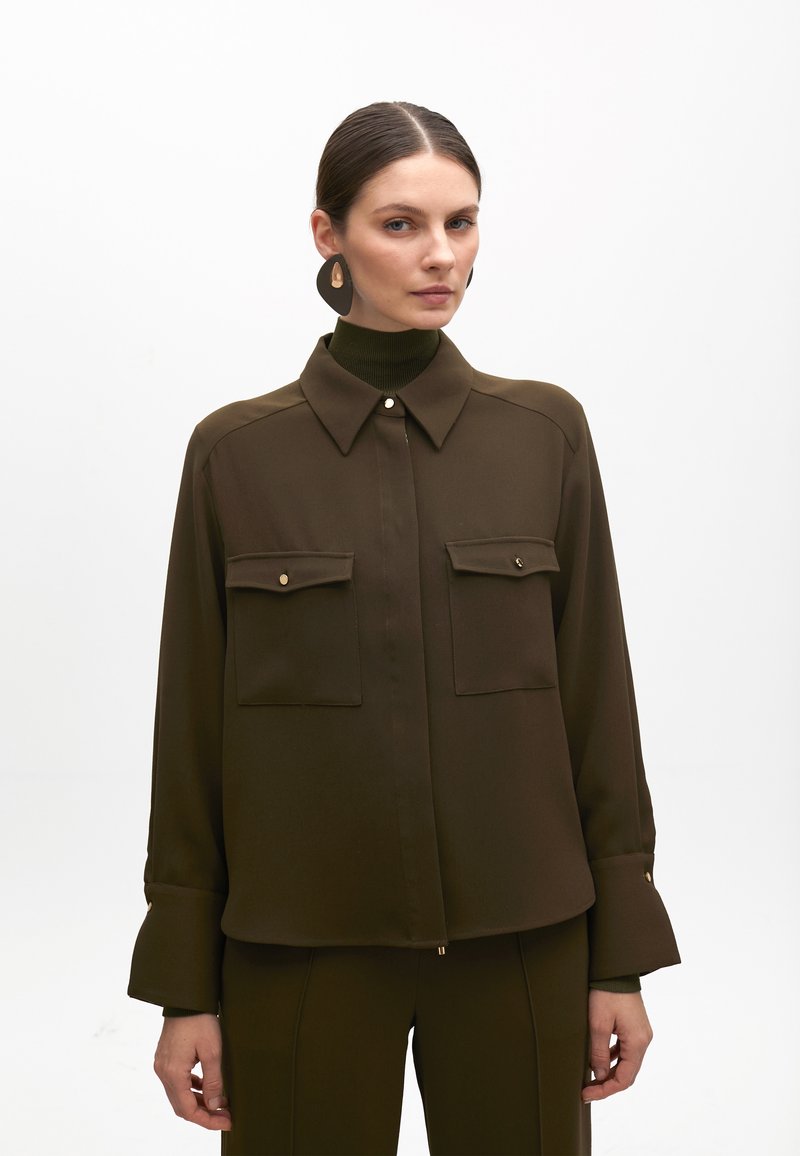 Blusa marrone a maniche lunghe con colletto alto, due tasche frontali, dettagli in bottone color oro e tessuto testurizzato, abbinata a un dolcevita coordinato.