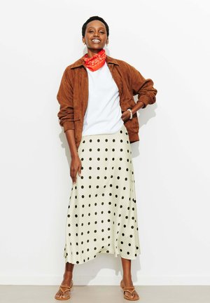 Femme souriante, portant une veste marron, une chemise blanche, une jupe crème à pois noirs, un foulard orange autour du cou et des sandales, debout contre un mur blanc.