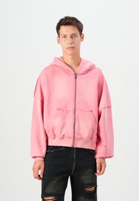 Lyserød cropped hoodie med lynlås foran, hætte, slidt kant og to forreste lommer. Matchet med sorte slidte jeans.