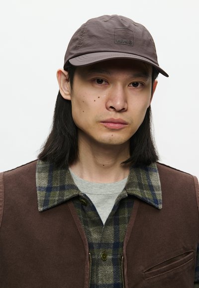 Pilkas kepurė su logotipu, dėvima asmens su ilgais juodais plaukais, uždengta rudais liemenėmis ir languota marškinėlių, kurių spalvos - žalia, mėlyna ir pilka.